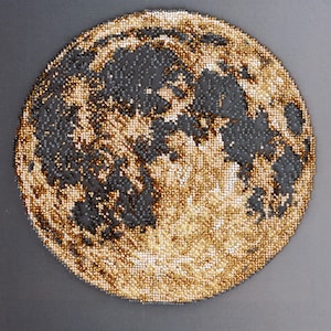 May include: Circular artwork depicting the moon, rendered in a mosaic style. The moon's surface is a mix of gold and black beads, creating a textured effect. The artwork is set against a gray background, enhancing the contrast and detail of the moon.