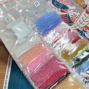 May include: A close-up of a variety of small, colourful beads in individual plastic bags. The bags are labelled with numbers, including 1, 17, and 19. There is also a spool of white thread in a separate bag.