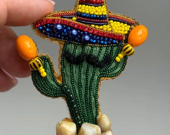 Könnte beinhalten: Handgefertigte Brosche mit einem grünen Kaktus, der einen Sombrero und einen schwarzen Schnurrbart trägt. Der Sombrero ist mit gelben, roten und blauen Perlen verziert. Der Kaktus hält orangefarbene Maracas und hat beige Steine an der Basis.