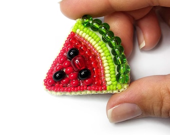 Könnte beinhalten: Eine Brosche in Form eines Wassermelonenstücks aus kleinen Perlen. Das Design zeigt eine dunkelgrüne äußere Schale, eine hellgrüne innere Schale und ein rotes Zentrum, das mit roten Perlen und drei schwarzen Perlen als Samen besetzt ist.