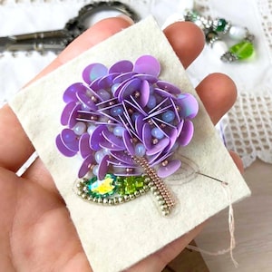 Puede incluir: Un broche hecho a mano con una flor de hortensia de lentejuelas moradas con un tallo de cuentas y una joya verde y amarilla. La flor está fijada a un cuadrado de tela beige, mostrando los intrincados detalles del diseño floral.