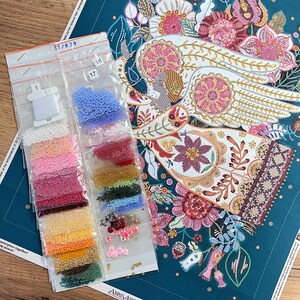 May include: A close-up of a colourful beaded embroidery kit featuring a design of a bird with outstretched wings. The kit includes a variety of beads in different colours and sizes, as well as a needle and thread. The beads are organised in small plastic bags labelled with numbers.