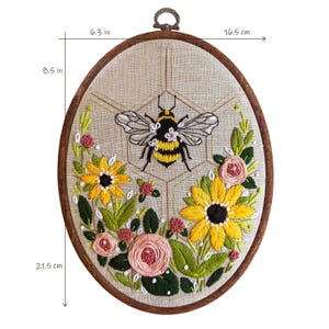 Könnte beinhalten: Stickerei in einem runden Rahmen mit einer gelben und schwarzen Hummel in einem Wabenmuster, umgeben von rosa und gelben Blumen. Der Rahmen hat einen Durchmesser von 21,5 cm.