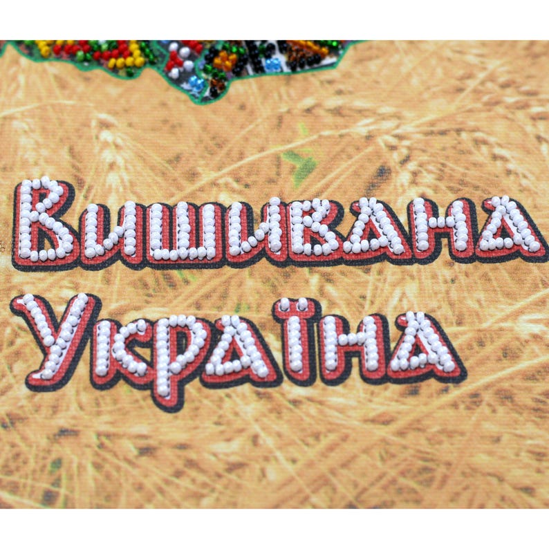 May include: Close-up of a canvas with the words "Вишивана Україна" in white beaded letters outlined in red. The background has a wheat field design. The top of the canvas has a colorful beaded design.