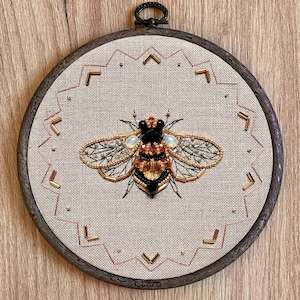 May include: Embroidered bee with gold and black details on a beige linen background. The bee is surrounded by a geometric pattern of gold thread. The embroidery is framed in a dark wood hoop.