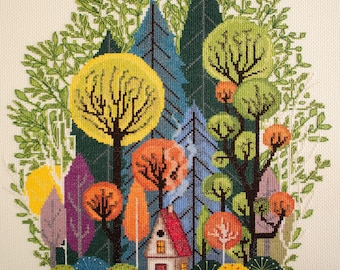 May include: A cross-stitch embroidery featuring a small house with a red roof nestled in a dense forest. The scene includes stylized trees in shades of yellow, orange, green, blue, and purple, with a light-colored path leading to the house entrance.
