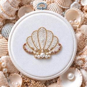 May include: An embroidered seashell design on a white fabric hoop. The shell is detailed with white beads, gold embellishments, and pearls. The background is scattered with seashells and pearls, evoking a seaside theme.