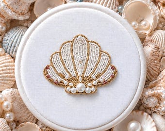 Könnte beinhalten: Ein weißer Stoff-Stickrahmen mit einem Muscheldesign. Die Muschel ist mit weißen Perlen, goldenen Akzenten und Perlen verziert. Der Hintergrund ist mit Muscheln und Perlen gefüllt, was ein Küstenmotiv erzeugt.