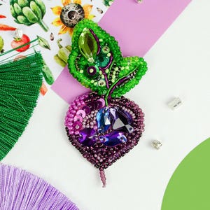 May include: A handcrafted beet brooch featuring deep purple and magenta seed beads, sequins, and faceted crystals. The leafy top is composed of vibrant green seed beads, sequins, and a central marquise-cut green crystal, creating a textured design.