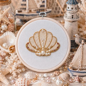 Puede incluir: Un bastidor de bordado circular de madera que contiene un panel de tela blanca con un diseño de concha marina cosido a mano. La concha está intrincadamente elaborada con cuentas de color crema, cuentas metálicas doradas y perlas blancas.