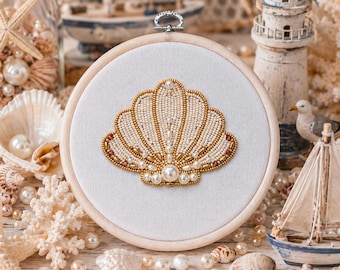 May include: A circular wooden embroidery hoop containing a white fabric panel with a hand-stitched seashell design. The shell is intricately crafted using cream-colored seed beads, gold-toned metallic beads, and various sizes of white pearls.
