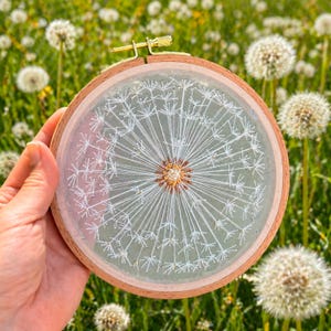 May include: A hand-stitched embroidery hoop featuring a detailed white dandelion design. The artwork is set against a pale green background and framed by a natural wood hoop. The dandelion's delicate seeds radiate from a central point.