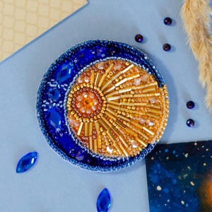 May include: A celestial-themed brooch featuring a crescent moon and sun design. The brooch is embellished with blue and gold beads, and blue crystal accents. The sun is in the center, with gold bead rays. The moon is on the left side, with blue crystal accents.