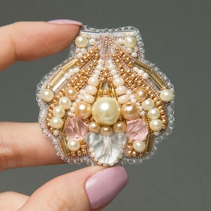 Könnte beinhalten: Muschelförmige Brosche, aufwendig mit cremefarbenen, goldenen und pfirsichfarbenen Perlen bestickt. Das Design umfasst klare und rosa Kristallakzente, wodurch ein schimmerndes, elegantes Accessoire entsteht.
