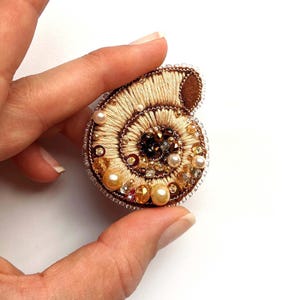 May include: A beige and brown embroidered brooch in the shape of a nautilus shell. The shell is decorated with pearls, beads, and sequins.
