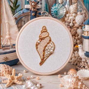 Puede incluir: Un bastidor de bordado circular de madera que contiene un panel de tela blanca con un diseño de concha marina cosido a mano. La concha está elaborada intrincadamente con una variedad de cuentas color crema, beige y blancas, perlas y oro.