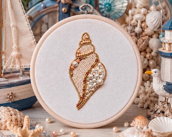 May include: A circular wooden embroidery hoop containing a white fabric panel with a hand-stitched seashell design. The shell is intricately crafted using a variety of cream, beige, and white beads, pearls, and metallic gold accents in a spiral pattern.