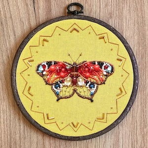 May include: A handcrafted, beaded butterfly art piece. The butterfly features red, orange, and blue beads on a yellow background, framed in a dark wooden hoop. The design includes a gold-colored stitched border and a metal hanging loop.
