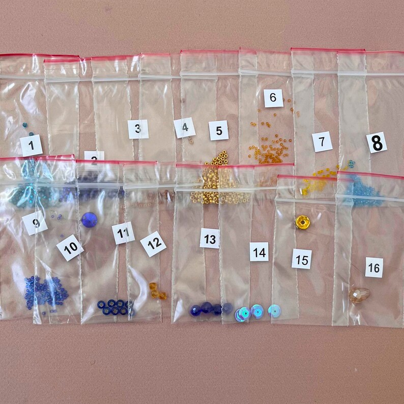 Pode incluir: Dezesseis pequenas sacolas de pl&aacute;stico etiquetadas com os n&uacute;meros de 1 a 16, cada uma contendo diferentes tipos de contas e lantejoulas. As sacolas est&atilde;o dispostas em quatro filas de quatro sacolas cada.