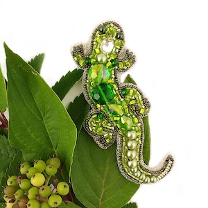 Puede incluir: Un broche de lagarto verde con cuentas con detalles de perlas verdes y blancas. El broche está sobre una hoja verde.