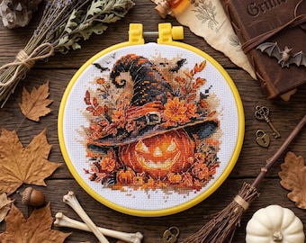 May include: A cross-stitch embroidery in a yellow plastic hoop featuring a jack-o'-lantern wearing a witch's hat, surrounded by autumn leaves and small bats. The design is stitched on white fabric with vibrant orange, black, and gold thread.