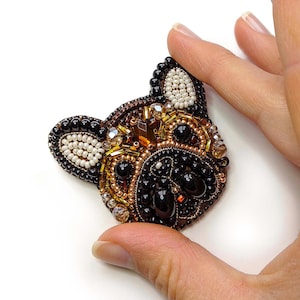 Può includere: Un broche di perline che rappresenta il viso di un bulldog francese. Il broche è realizzato con perline nere, marroni, dorate e bianche. Gli occhi del cane sono fatti di perline nere e il naso è fatto di una perlina marrone.