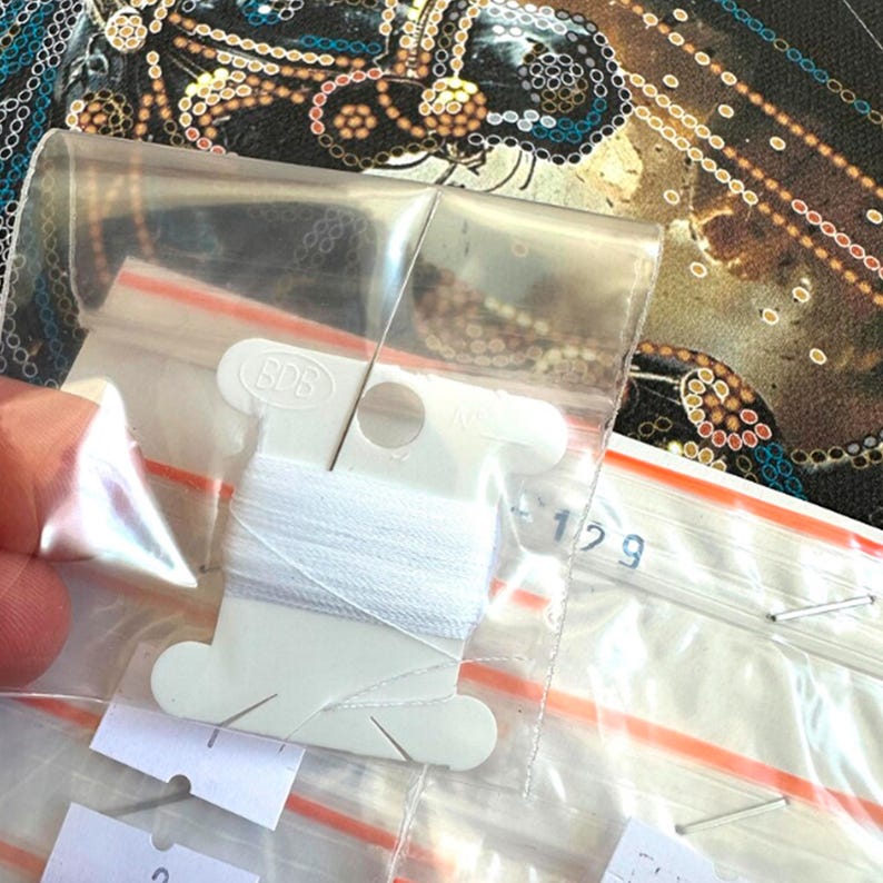 Puede incluir: Primer plano de una bobina de hilo blanco con una aguja, dentro de una bolsa de pl&aacute;stico transparente. La bolsa est&aacute; parcialmente abierta, revelando el hilo y una peque&ntilde;a etiqueta. El fondo presenta una tela estampada con colores oro, azul y negro.
