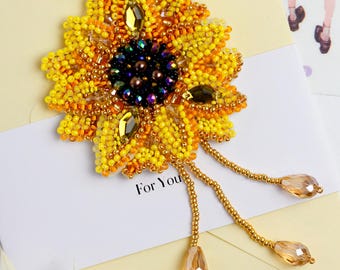 May include: A beaded sunflower brooch with a yellow and orange flower and a dark brown center. The brooch has three strands of beads hanging from the bottom, each ending in a clear teardrop-shaped bead. The text "For You" is written on a white card behind the brooch.