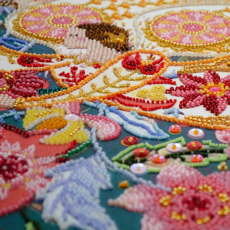 May include: Close-up of a colourful beaded embroidery design featuring flowers, leaves, and swirls. The beads are arranged in a variety of colours, including red, pink, yellow, blue, and green. The embroidery is done on a white fabric background.