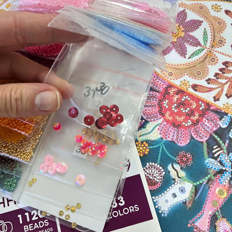 May include: Close-up of small plastic bags filled with colourful beads. The bags are labelled with numbers, including "3130" and "32". The beads are in various colours, including red, pink, and gold. The bags are on a table with a larger image of a colourful floral design.