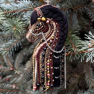May include: A handcrafted horse head ornament with a detailed design. The horse features a black braided mane, brown and gold accents, and beaded embellishments. Silver chains and dangling beads add to the decorative appeal. The ornament is suspended against a backdrop of evergreen branches.