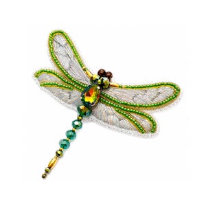 Peut inclure: Une broche libellule ornée de perles de cristal vert et doré, de broderies en fil métallique argenté sur les ailes et d'une pierre centrale en forme de goutte aux reflets multicolores. Les ailes sont bordées de petites perles vertes et blanches.