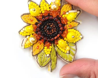 May include: A yellow and orange embroidered sunflower brooch with a beaded center. The sunflower has a green stem and leaves.