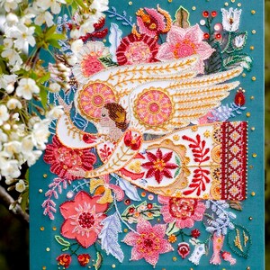 May include: A colourful beaded artwork featuring a white bird with outstretched wings, surrounded by flowers and leaves. The artwork is set against a teal background.