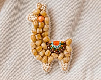 May include: A llama-shaped brooch crafted with tan and beige stone beads, accented by a bright orange bead for the eye and a saddle design featuring turquoise, orange, and metallic brown beads, all outlined with small white seed beads.