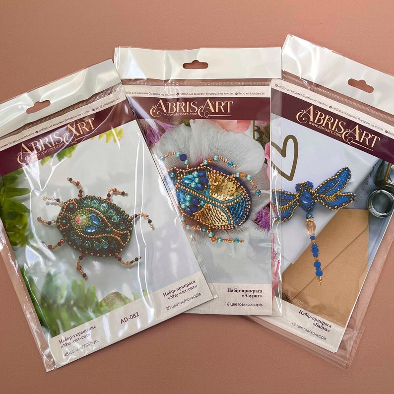 Pode incluir: Tr&ecirc;s pacotes de kits de bordado com contas que apresentam um besouro azul e dourado, uma lib&eacute;lula azul e um besouro azul e verde. Cada pacote inclui as contas, o fio e as instru&ccedil;&otilde;es para criar o design. Os pacotes s&atilde;o etiquetados com o nome "Abris Art" e o endere&ccedil;o do site "www.abrisart.com".