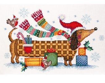 May include: A cross-stitch pattern featuring a brown dachshund wearing a red Santa hat and a long, multicolored striped scarf. The dog is surrounded by wrapped Christmas gifts, two small birds, and light blue snowflakes on a white fabric background.