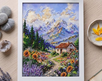 May include: A framed diamond painting featuring a landscape with a small house, a winding path, sunflowers, and lavender in the foreground, set against a backdrop of green pine trees and snow-capped mountains under a blue sky with clouds.