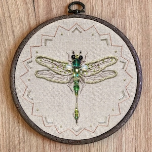 May include: A green dragonfly embroidered with beads and sequins in a circular wooden embroidery hoop. The dragonfly has a long, beaded tail and its wings are detailed with brown thread.