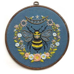 May include: Embroidered hoop art featuring a bee with yellow and black stripes surrounded by a wreath of flowers on a blue background.