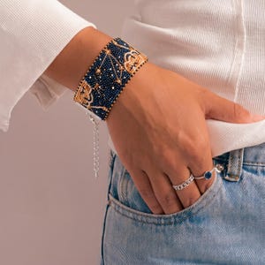 May include: A wide cuff bracelet crafted from dark blue and gold seed beads featuring a repeating geometric pattern. The bracelet includes a silver-toned metal extender chain with a lobster claw clasp. Two silver rings are also visible on the fingers.