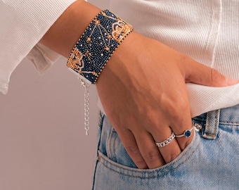 May include: A wide cuff bracelet crafted from dark blue and gold seed beads featuring a repeating geometric pattern. The bracelet includes a silver-toned metal extender chain with a lobster claw clasp. Two silver rings are also visible on the fingers.