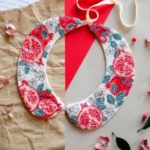 May include: A white fabric collar with a red and green embroidered design of pomegranates. The collar is tied with a white ribbon.