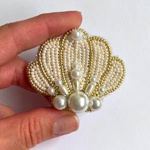 Puede incluir: Un broche con forma de concha marina elaborado con intrincado trabajo de cuentas. El diseño presenta filas alternas de cuentas de color crema y oro metálico, acentuadas con perlas artificiales blancas de varios tamaños en abanico.