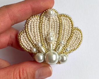 Könnte beinhalten: Eine muschelförmige Brosche aus aufwendigen Perlenstickereien. Das Design besteht aus abwechselnden Reihen von cremefarbenen und goldfarbenen Saatperlen, akzentuiert durch verschieden große, glatte weiße Kunstperlen in Fächerform.
