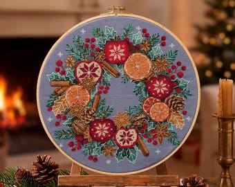 May include: A circular embroidery hoop containing a cross-stitch pattern of a holiday wreath on light blue fabric. The wreath features red apples with white snowflake designs, orange slices, cinnamon sticks, pinecones, and green holly leaves.