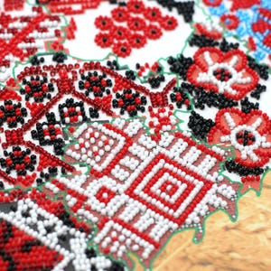 May include: Close-up of a detailed, beaded embroidery with a traditional floral and geometric design. The pattern features red, white, and black beads on a white background, with green outlining. The design is reminiscent of folk art.