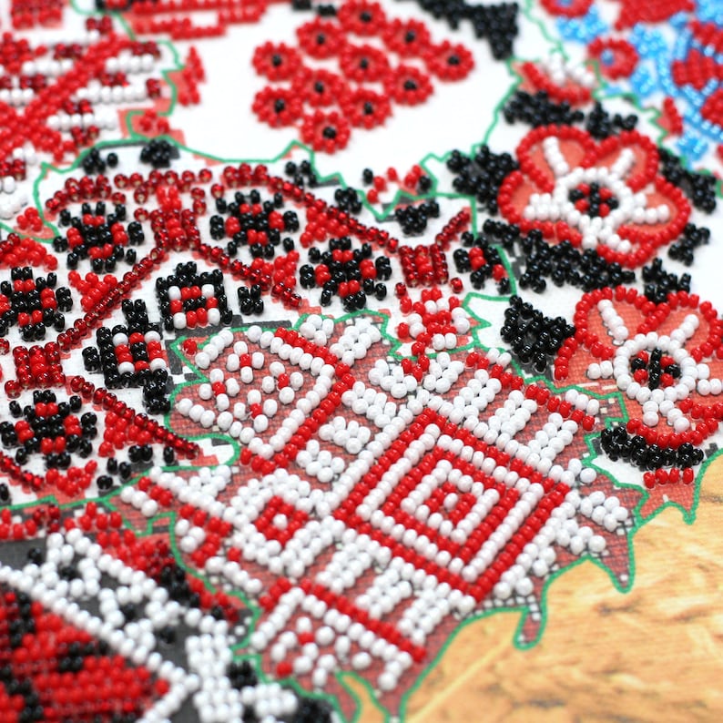 May include: Close-up of a detailed, beaded embroidery with a traditional floral and geometric design. The pattern features red, white, and black beads on a white background, with green outlining. The design is reminiscent of folk art.