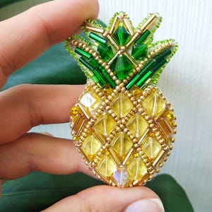 以下が含まれることがあります： 黄色、緑、金色のビーズで作られた手作りのパイナップルのブローチ。ブローチは、黄色のボディにダイヤモンド模様、緑の王冠と、詳細なデザインが特徴です。トロピカルなアクセサリーとして最適です。