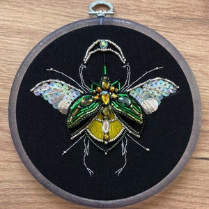 May include: A green and gold beetle with iridescent wings is embroidered with beads and sequins on black fabric in a circular wooden frame.